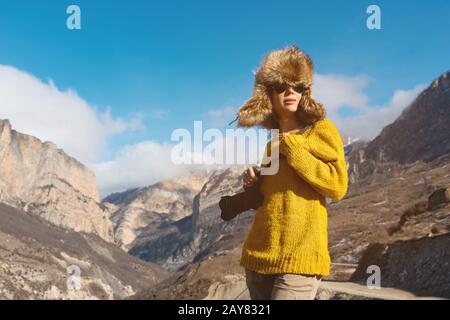 Ein Girl-Fotograf in Sonnenbrille und ein großer Fellhut und ein gelber Strickpullover stehen vor dem Hintergrund hoher Felsen in Stockfoto