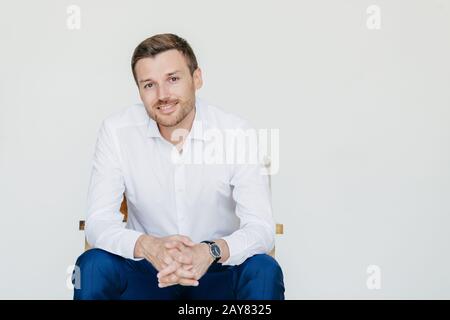 Studio mit elegantem, fröhlichem männlichen Unternehmer in formeller Kleidung, sitzt auf dem Stuhl, posiert an der Kamera, isoliert über weißem Hintergrund. Froher Mann lächelt h Stockfoto