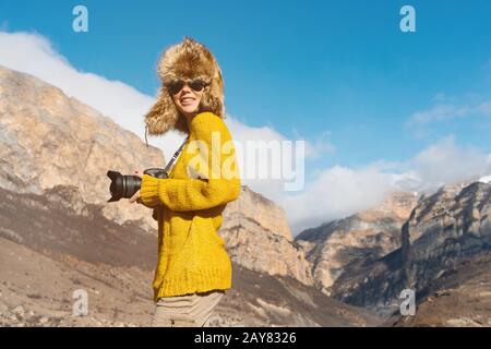 Ein Girl-Fotograf in Sonnenbrille und ein großer Fellhut und ein gelber Strickpullover stehen vor dem Hintergrund hoher Felsen in Stockfoto