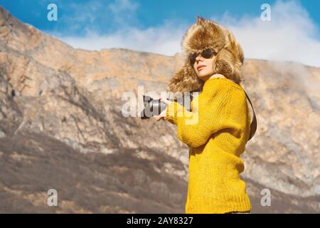 Ein Girl-Fotograf in Sonnenbrille und ein großer Fellhut und ein gelber Strickpullover stehen vor dem Hintergrund hoher Felsen in Stockfoto