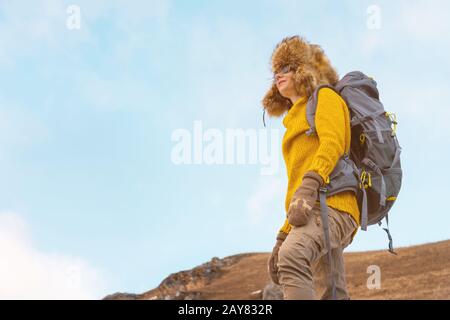 Das Backpacker-Mädchen in Sonnenbrille und ein großer Nordfellhut mit einem Rucksack auf dem Rücken steht auf einem Felsen und sieht towar aus Stockfoto