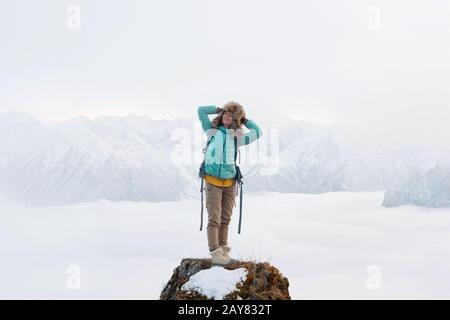 Ein fröhliches lächelndes Mädchen aus dem Flusspferd reist in einer Daunenjacke mit Rucksack und steht in einem großen Fellhut auf einem Felsen Stockfoto