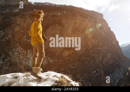 Ein Girl-Fotograf in Sonnenbrille und ein großer Fellhut und ein gelber Strickpullover stehen vor dem Hintergrund hoher Felsen in Stockfoto