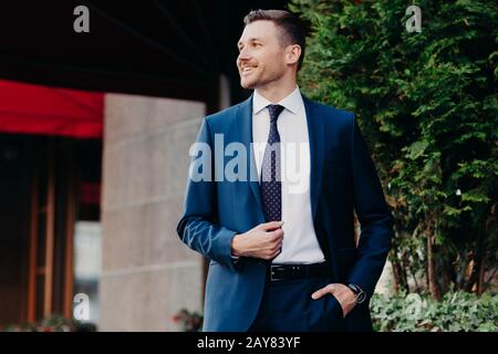 Wohlhabender gutaussehender Geschäftsmann im formellen Anzug, hält Hand in der Tasche, sieht positiv beiseite, steht in der Nähe der Bank, hat fröhlichen Ausdruck, positive Schimper Stockfoto