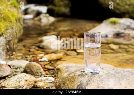 Auf einem Stein neben dem Gebirgsfluss steht ein transparentes Glasglas mit Mineralbergwasser Stockfoto