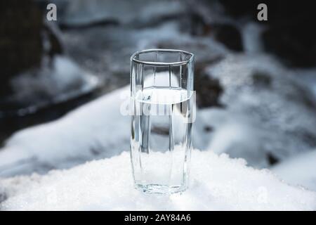 Im Schnee steht vor dem Hintergrund eines sauberen Frostbergsflusses i ein transparentes Glasglas mit Trinkwasser Stockfoto