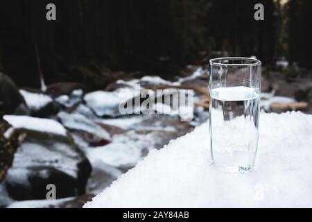 Im Schnee steht ein transparentes Glasglas mit Trinkwasser vor dem Hintergrund eines sauberen Gebirgsflusses und eines f Stockfoto