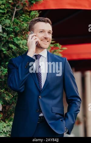 Vertikaler Schuss zufriedener männlicher Mitarbeiter hat Telefongespräch, in schwarzen Anzug gekleidet, hält Hand in Tasche, hat guten Ausdruck, sanftes sm Stockfoto