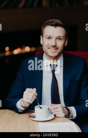 Zufriedener männlicher Mitarbeiter im Luxusanzug, Kaffee trinken, sitzt am Holztisch in der Cafeteria, hat ein angenehmes Lächeln und einen attraktiven Look, wartet auf Business p Stockfoto