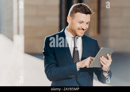 Erfolgreicher männlicher Unternehmer im formellen Anzug, hält modernes Tablet, prüft die Dokumentation online, liest die notwendigen Informationen für die Erstellung von Geschäftsberichten, Stockfoto