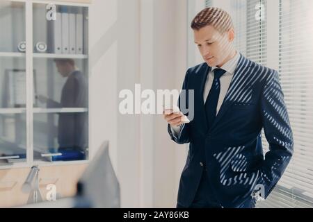 Attraktiver, in Unternehmenskleidung gekleideter Männerdirektor, checkt E-Mails auf Handy, steht in der Nähe von Fenster mit Verschluss in seinem gemütlichen Schrank, bloggt websi Stockfoto