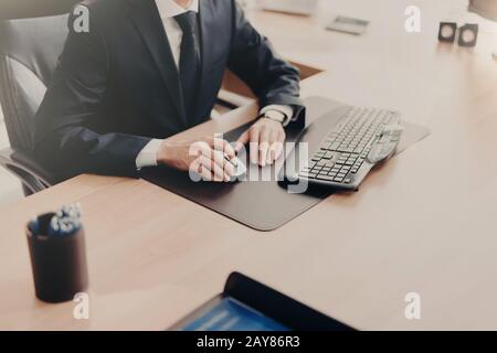 Zugeschnittenes Bild eines erfolgreichen männlichen Geschäftsinhabers sitzt am Arbeitsplatz in einem formellen Anzug, verwendet Computer für die Arbeit, erstellt einen Finanzbericht. Führungskraft mit männlichem Stockfoto