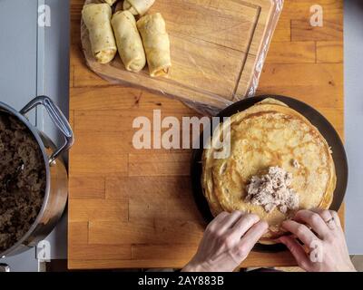 Draufsicht über menschliche Hände, die Fleisch in frisch gebackenen Pfannkuchen auf Holzgrund einwickeln. Gerollte Pfannkuchen mit Füllung werden neben dem Kitch gelegt Stockfoto