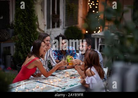 Fünf Erwachsene, die mit Cocktails toasten, an der Bar im Freien, lachen und Spaß haben. Spaß, Party, Geselligkeit, multiethnische, introvertierte und extrovertierte Perso Stockfoto