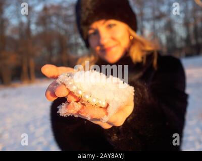 Eine junge Frau in einem schwarzen Hut und einem minken Fellmantel hält in ihren Handflächen eine Handvoll flauschigen weißen Schnee, in dem ein weißer Perlenschmuck liegt. Perlen und Sno Stockfoto