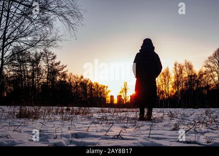 Silhouette einer weiblichen Figur, die im Winter in der Hintergrundbeleuchtung der untergehenden Sonne im Schnee steht. Eine Frau blickt auf die Sonne über städtischen Häusern hinter Th Stockfoto