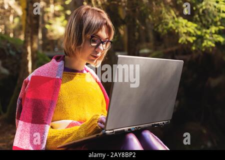 Ein gezungtes Porträt eines lächelnden freiberuflichen Flusspferdes mit einer Brille, die in einer Decke mit einem Laptop gekleidet ist, der auf einem Rost sitzt Stockfoto