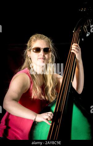 Weibliche Musikerin mit rotem Hemd spielt auf einem Kontrabass vor schwarzem Hintergrund Stockfoto