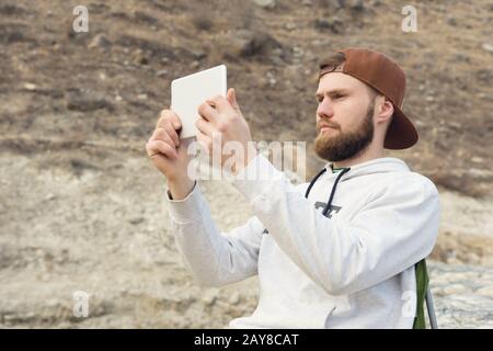 Mit einem Bart in einer Mütze und einem Sweatshirt mit einem Tablett in den Händen fotografiert der Hipster im Freien auf einem Tablet Stockfoto
