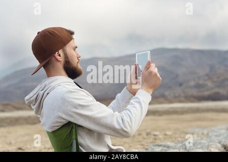 Mit einem Bart in einer Mütze und einem Sweatshirt mit einem Tablett in den Händen fotografiert Hipster auf einem Tablett, während er auf der Natur ist B. Stockfoto