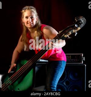 Weibliche Musikerin mit rotem Hemd spielt auf einem Kontrabass vor schwarzem Hintergrund Stockfoto