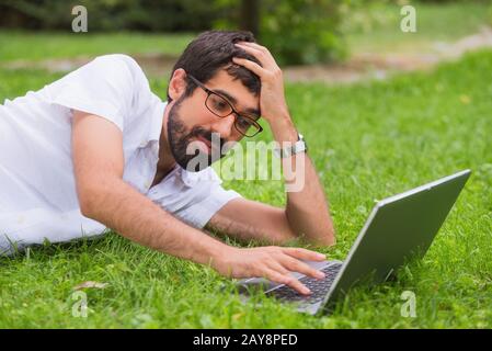 Junger Mann bei der Arbeit auf seinem Laptop beim Liegen auf dem Rasen des Parks. gelangweilter Gesichtsausdruck. Stockfoto