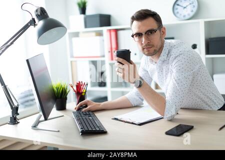 Ein junger Mann in Gläsern steht in der Nähe eine Tabelle im Büro, hält ein Glas Kaffee in der Hand und mit einem Computer funktioniert. Stockfoto