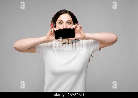 Portrait von emotionalen schöne Happy plus größe Modell in Kopfhörer holding Handy hinter ihren Mund hören von Musik Stockfoto