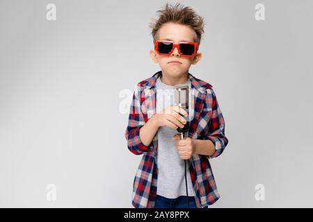 Ein gutaussehender Junge in einem Kraghemd, grauem Hemd und Jeans steht auf grauem Hintergrund. Ein Junge, der eine Sonnenbrille trägt. Der rothaarige Junge Stockfoto