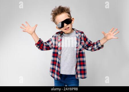 Ein gutaussehender Junge in einem Kraghemd, grauem Hemd und Jeans steht auf grauem Hintergrund. Der Junge in schwarzer Sonnenbrille. Der Junge breitete h aus Stockfoto