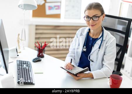 Ein junges Mädchen in einer weißen Robe sitzt an einem Tisch im Büro und hält einen Stift und ein Notizbuch in der Hand. Ein Stethoskop hängt aroun Stockfoto