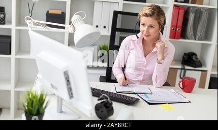 Ein junges Mädchen in der Kopfhörer sitzt an einem Tisch im Büro, hält einen Bleistift in der Hand und arbeitet mit einem Notebook und einem Computer. Stockfoto