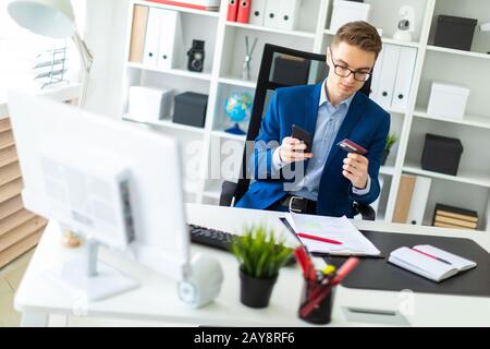 Ein junger Mann sitzt an einem Tisch im Büro und hält eine Bankkarte und Telefon. Stockfoto
