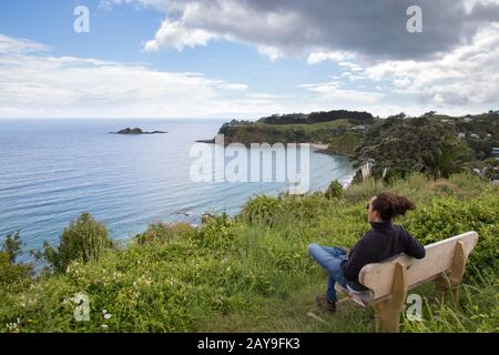 Männlich, auf Holzbank am Rand der Klippe sitzend, mit Blick auf den Ozean Stockfoto