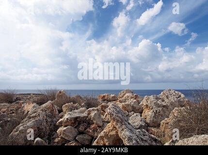 Raue Felsen und Felsbrocken mit trockener Sommervegetation vor einem ruhigen Meer mit weißen, sonnenbeleuchteten Wolken Stockfoto