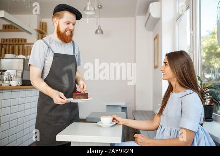 Kaffee-Business-Konzept - Kellner oder Barkeeper, der Schokoladenkuchen gibt und mit der kaukasischen schönen Dame in blauem Kleid bei C spricht Stockfoto