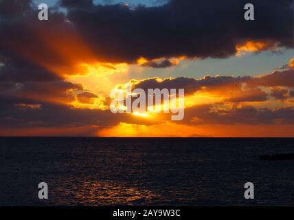 Sonnenuntergang über einem ruhigen, dunklen Meer mit orangefarbenen Strahlen, die durch dunkle dramatische Abendwolken glänzen Stockfoto