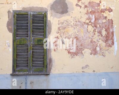 Eine alte gerissene Hauswand, die in abblätternden gelben und roten Farben und grünen Fensterläden gemalt wurde Stockfoto