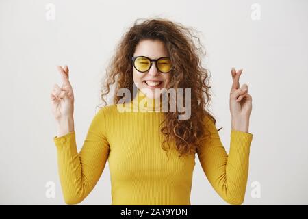 Aufgeregte Frau in gelbem Pullover, die die Finger durchzogen hält, Mund weit offen, wartet auf einen besonderen Moment, der auf grauem Hintergrund isoliert ist Stockfoto