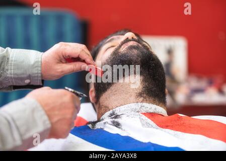 Barber rasiert den Bart eines gutaussehenden bärtigen Mannes mit einem elektrischen Rasierer im barber Shop. Stockfoto