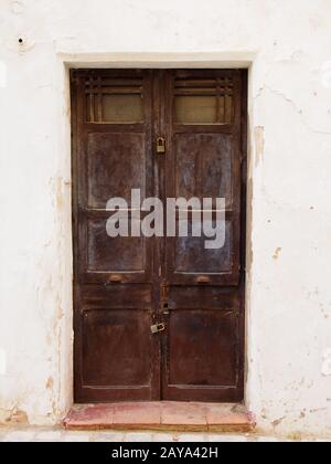 Alte, braune Doppeltüren aus Holz mit blätternd verblichener Schälfarbe und rostigen Griffen, mit einem Vorhängeschloss verschlossen Stockfoto