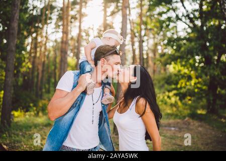 Familien-Mutter, Papa und Tochter sitzen am Papa auf den Schultern, und Eltern küssen sich im Sommer im Wald um die Natur Stockfoto