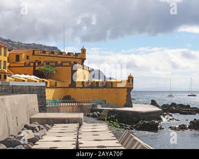 Blick auf das gelbe Fort von São Tiago aus dem 17. Jahrhundert in funchal madeira, von der Küste mit sonnenbeleuchteten blauen Meer und Boot Stockfoto