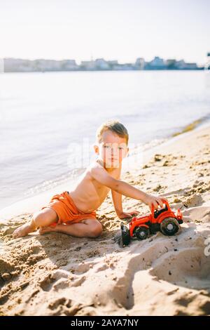 Thema Bauwesen und Schwerindustrie. Abstraction Kinderjunge, die im Sommer auf dem Sand in der Nähe des Flusses spielen, Toy Red Traktor Stockfoto