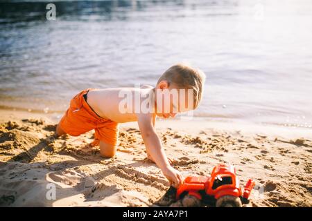Thema Bauwesen und Schwerindustrie. Abstraction Kinderjunge, die im Sommer auf dem Sand in der Nähe des Flusses spielen, Toy Red Traktor Stockfoto