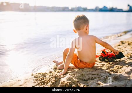 Thema Bauwesen und Schwerindustrie. Abstraction Kinderjunge, die im Sommer auf dem Sand in der Nähe des Flusses spielen, Toy Red Traktor Stockfoto