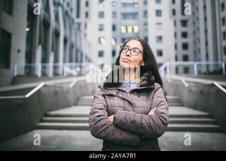 Schöne junge Frau europäischer Ethnie mit langen brünettigen Haaren, die nach oben und links schauen, Brille und Mantel tragen, steht auf B. Stockfoto