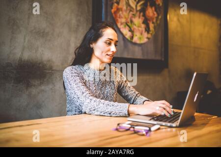 Schöne junge Brunette-Frau, die einen Laptop im Café an einem Holztisch in der Nähe eines Fensters verwendet, um Text auf einer Tastatur einzugeben. Im Winter Stockfoto