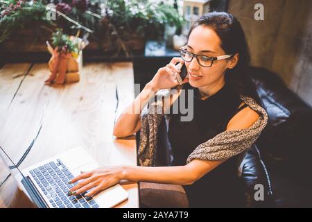 Schöne junge Brunette-Frau im Café mit weihnachtseinrichtung am Holztisch am Fenster telefonieren und sehen Stockfoto