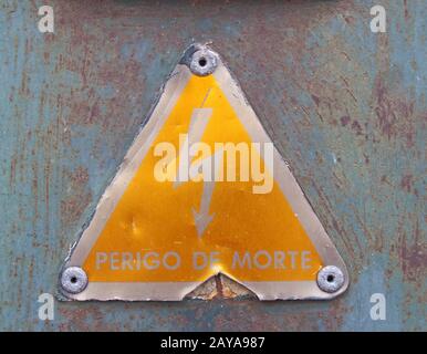 Ein altes dreikantiges gelbes, gelbes Portugiesisch-elektrisches Sicherheitszeichen aus Metall mit einem blitzkräftigen Symbol, das Perigo de morte - Transla - liest Stockfoto